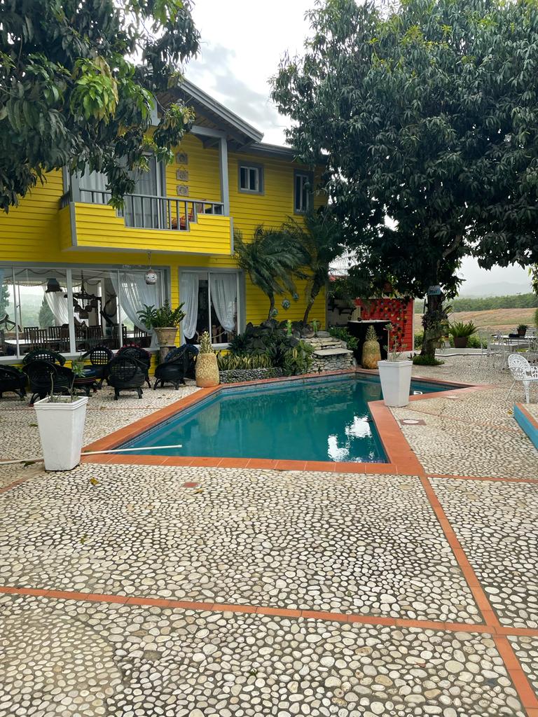 A colorful two-story house is framed by lush greenery and features ample outdoor seating. A serene pool is visible in the foreground, surrounded by textured stone pathways. Natural light is enhanced by the openness of the space, inviting relaxation and leisure.