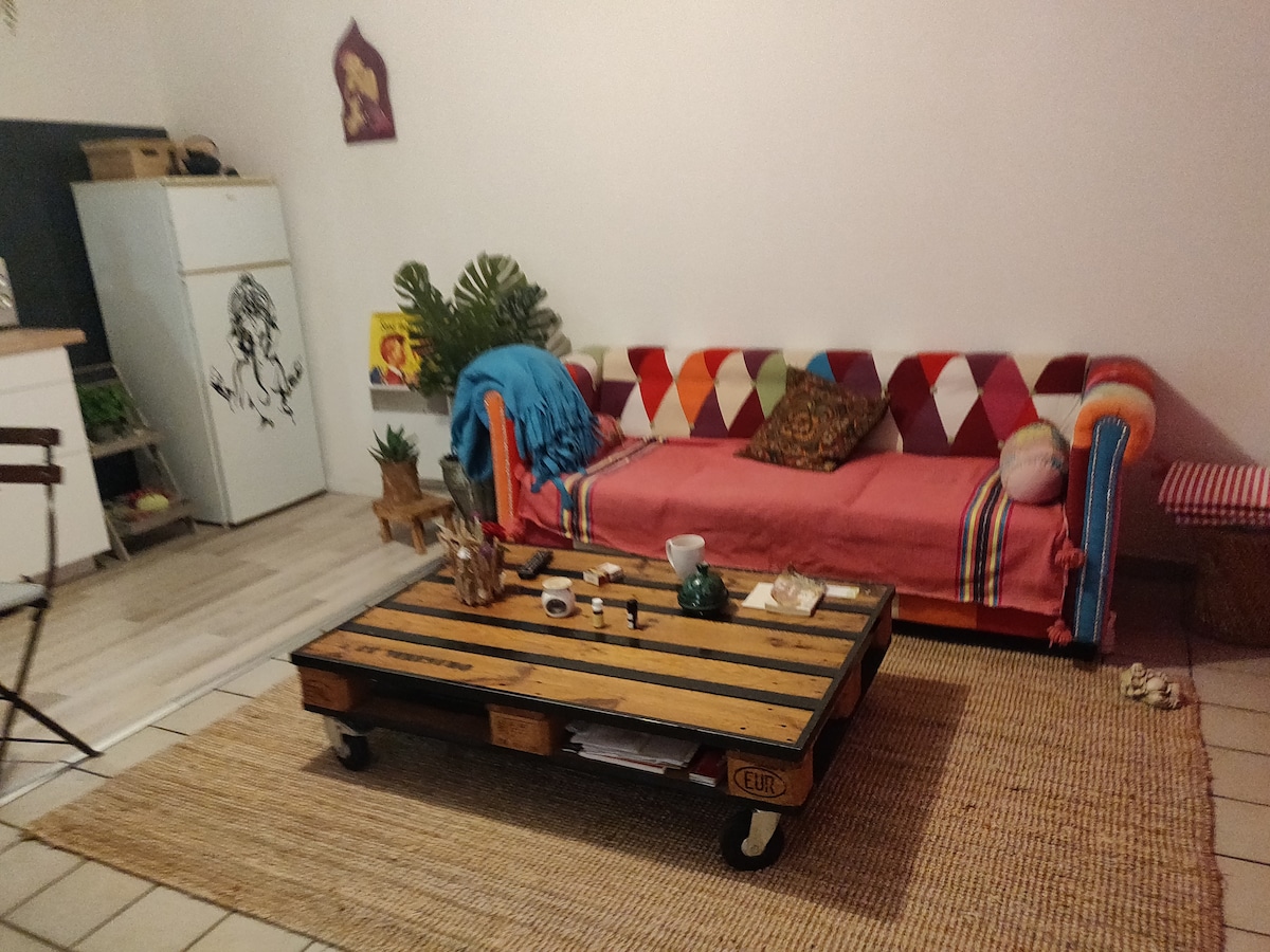 A colorful living area features a patchwork sofa adorned with cushions and a woven throw. A rustic coffee table sits on a jute rug, accompanied by decorative items. A kitchen area is visible in the background, complemented by a potted plant adding a touch of greenery.