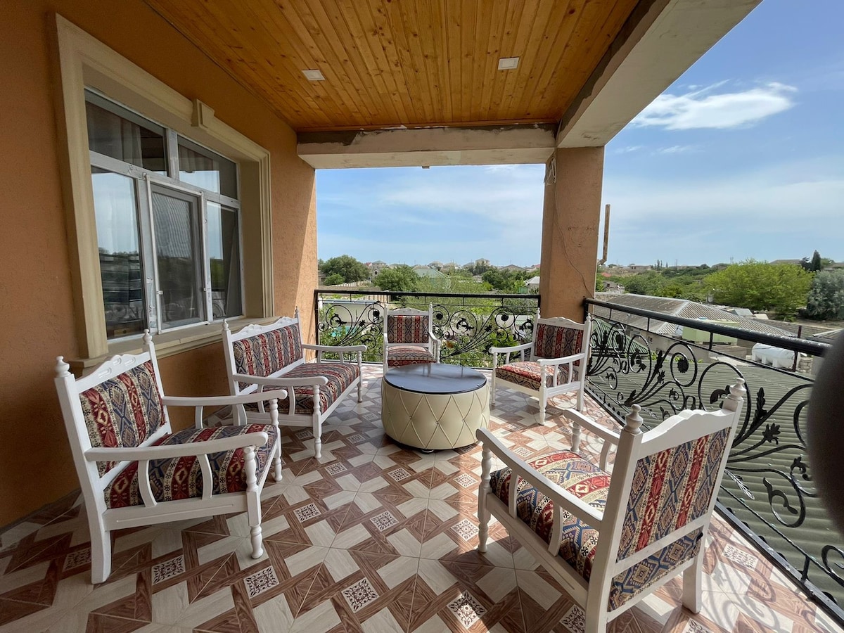A balcony is presented with a wrought-iron railing overlooking green surroundings. Five white chairs with patterned cushions circle a round table, inviting relaxation. Natural light fills the space through a large window, complementing the wooden ceiling and tiled floor.