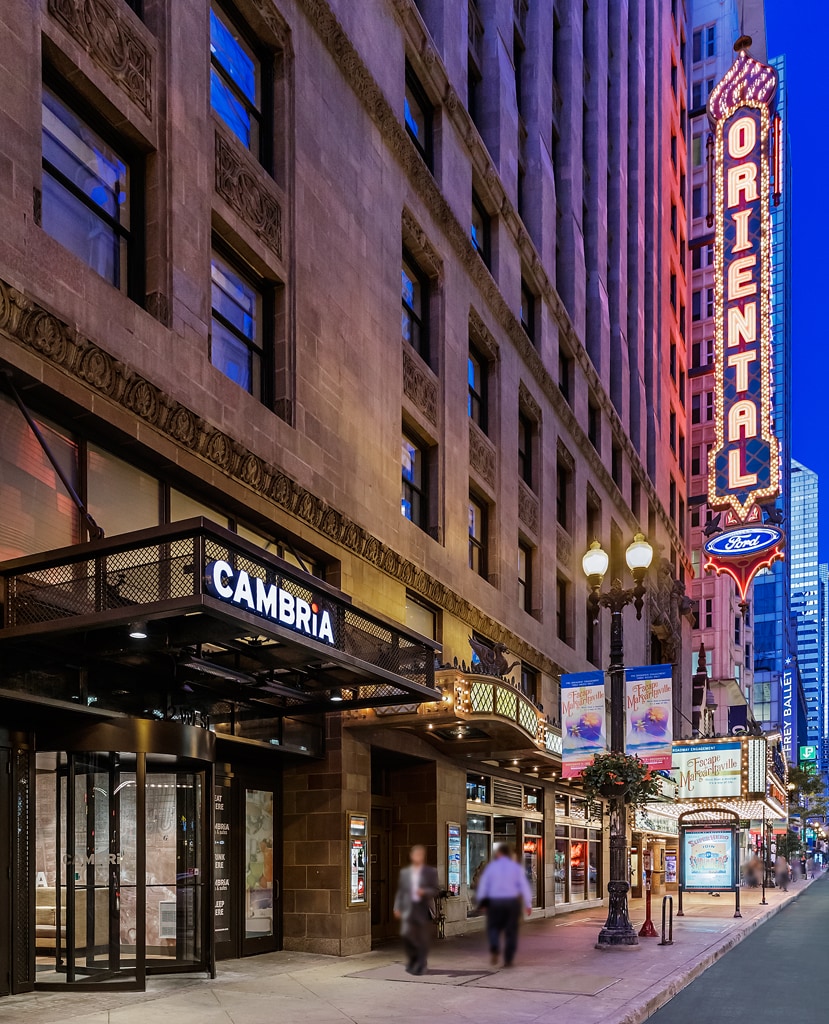 The hotel entrance features a modern black awning with the Cambria logo prominently displayed. Flanking the entrance are elegant decorative elements and bright signage from the adjacent Oriental Theatre, contributing to a vibrant urban atmosphere.