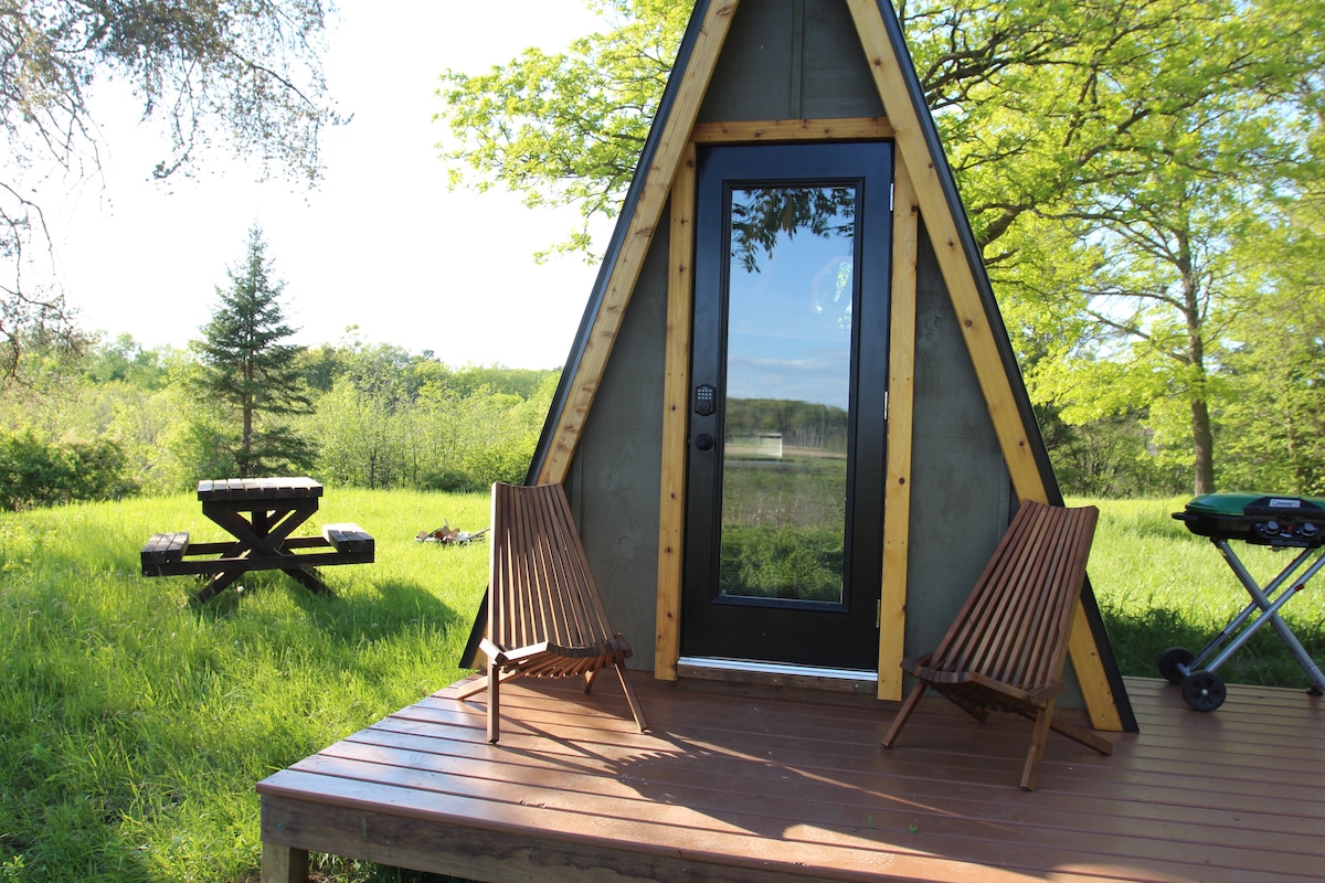 A rustic a-frame structure is featured, set against a backdrop of lush greenery. A wooden deck hosts two Adirondack chairs, inviting relaxation. A picnic table is positioned nearby, while a grill is placed to the side, completing this outdoor setting with nature surrounding the cabin.