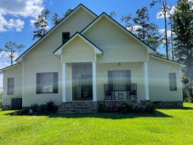 A welcoming house is depicted, featuring a light-colored exterior with a green accent roof. Rocking chairs are placed on the front porch, and a landscaped garden is visible in front. Large windows allow natural light into the home, surrounded by a grassy area and trees.