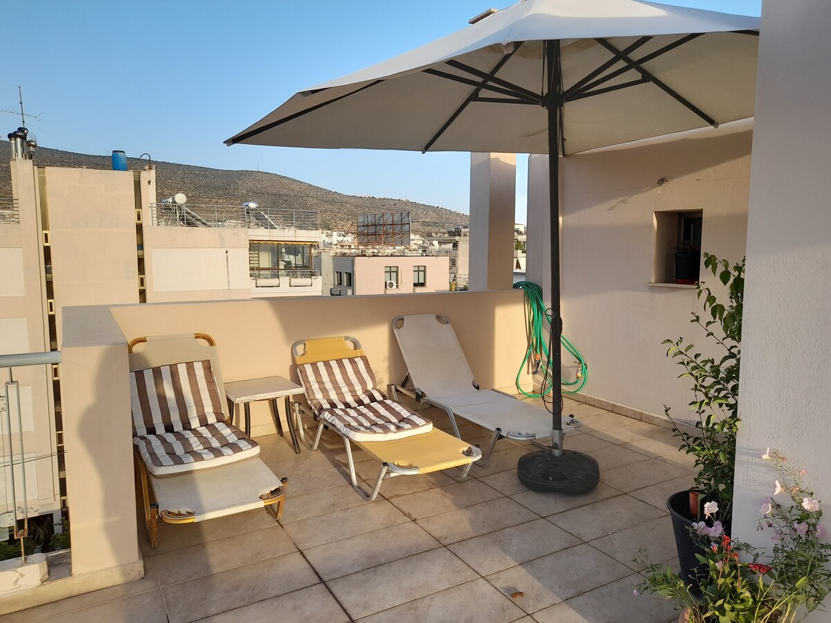 A sunlit rooftop terrace features two lounging chairs with striped fabric and a large umbrella providing shade. A green hose is coiled neatly nearby, and a potted plant adds a touch of greenery to the space. The surrounding buildings and hills are visible in the background.