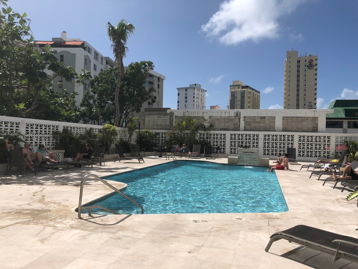 A private pool is framed by palm trees and lounge chairs, providing a relaxing outdoor space. The clear blue water reflects sunlight, while the surrounding area features well-maintained landscaping. High-rise buildings are visible in the background, showcasing the urban setting.