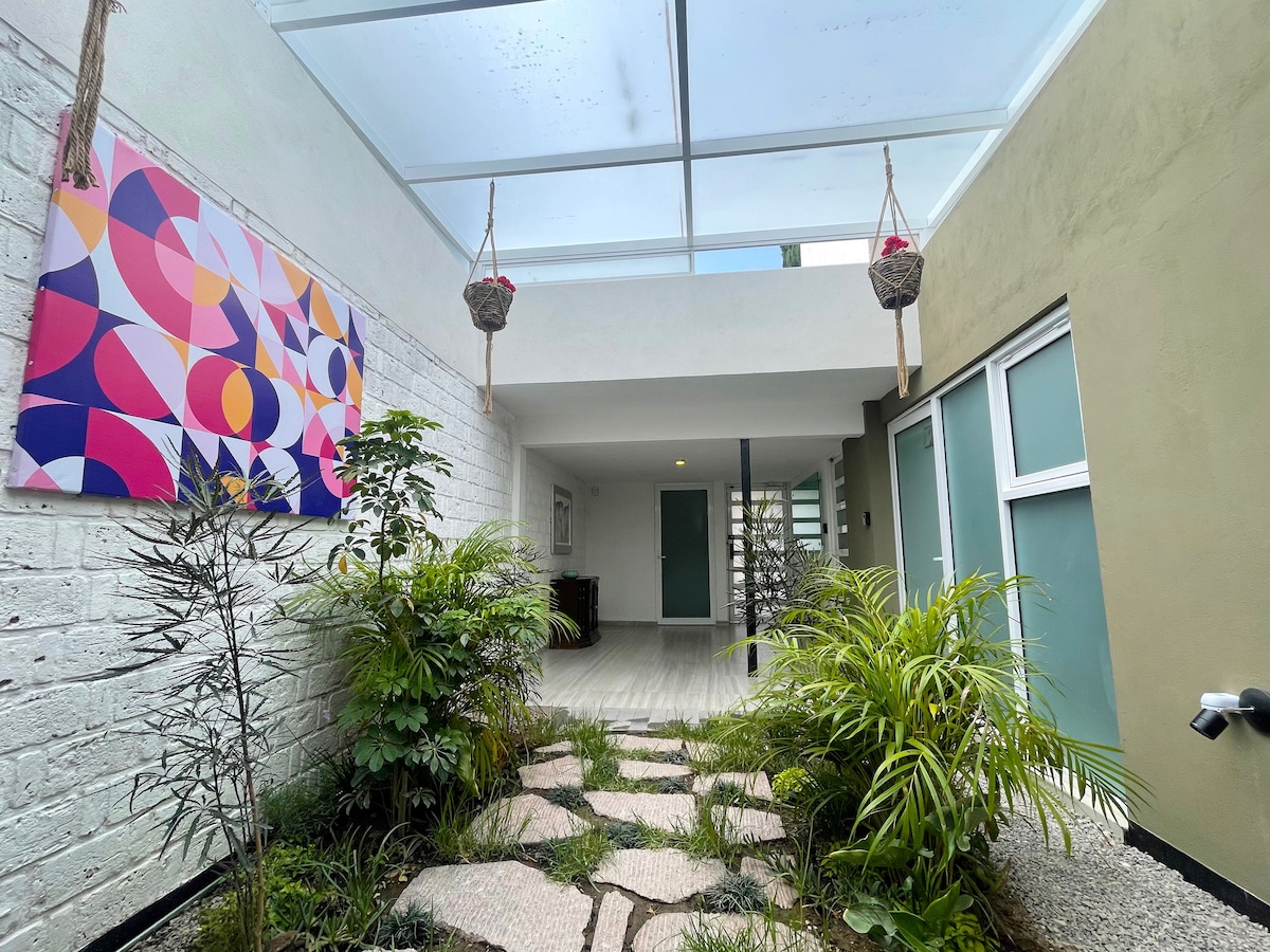 A bright indoor walkway is illuminated by a glass ceiling, featuring a mix of greenery and decorative plants. Colorful artwork adorns one wall, while stone pathways lead towards the entrance of the lofts, creating an inviting atmosphere.