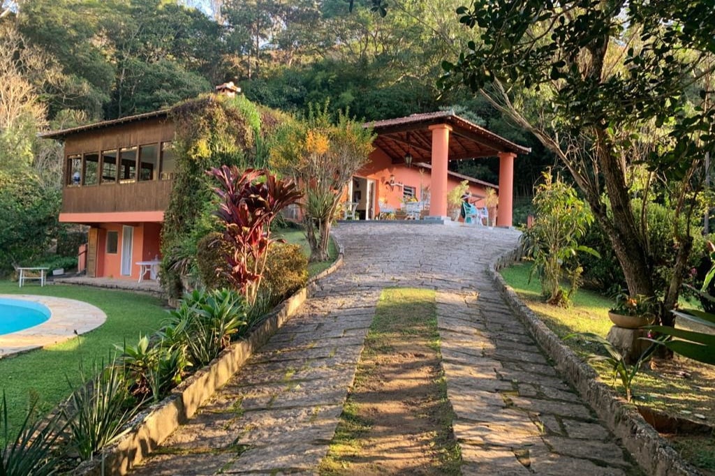 A winding stone path leads up to a vibrant orange house, surrounded by lush greenery. The spacious outdoor area features a glistening pool on the left, and tropical plants create a natural border along the path, enhancing the tranquil setting.