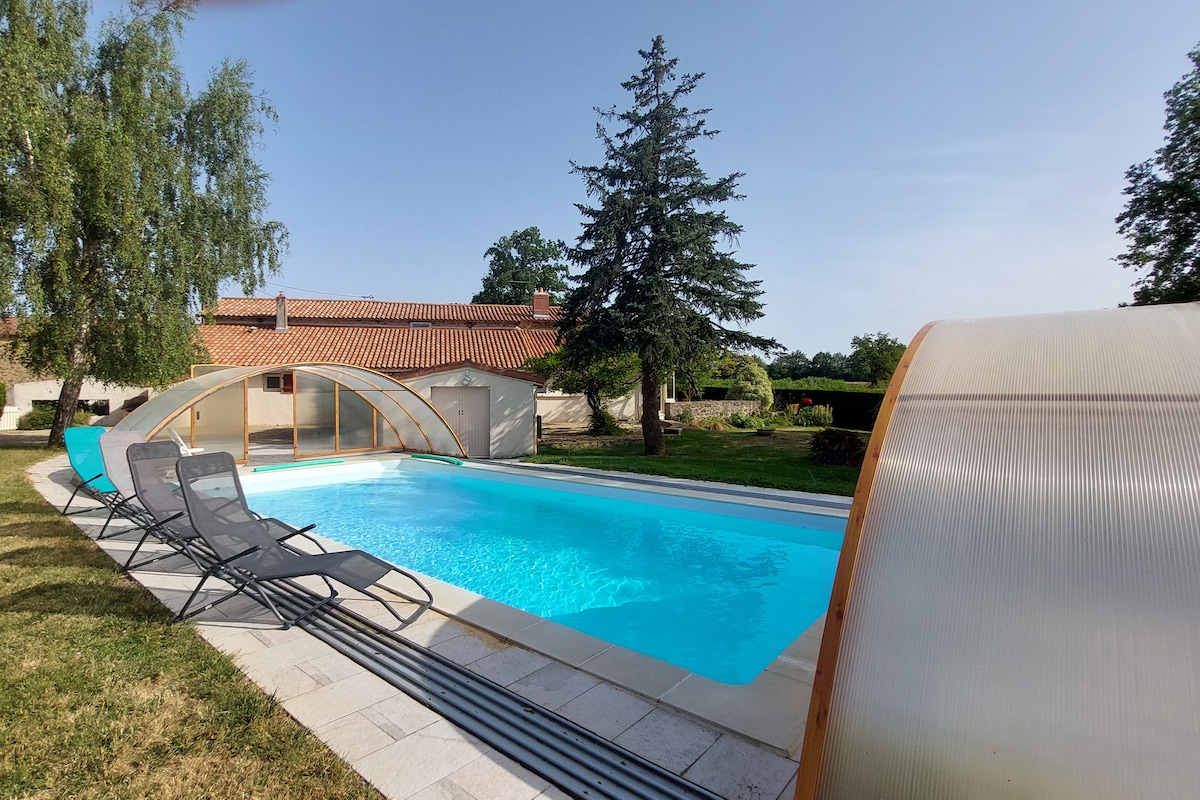 A private, covered swimming pool is situated within a landscaped garden, featuring lounge chairs arranged along the poolside. The pool's clear blue water contrasts with the surrounding greenery, while a gentle slope of lawn leads to the far end of the property.