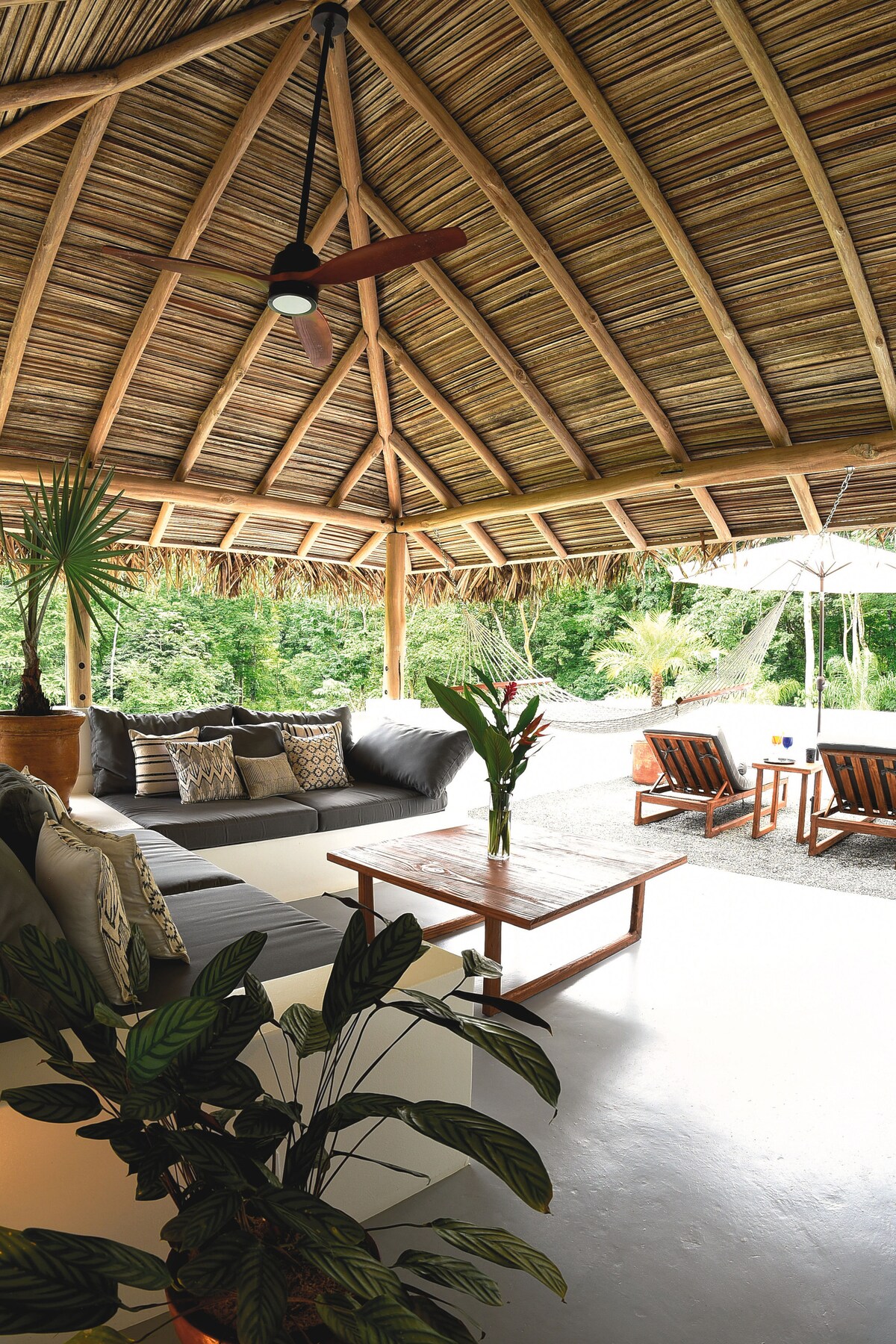 An open-concept outdoor lounge area features a thatched roof overhead and wooden beams. Comfortable seating with decorative pillows is arranged around a wooden coffee table. Lush greenery is visible in the background, enhancing the tranquil atmosphere.
