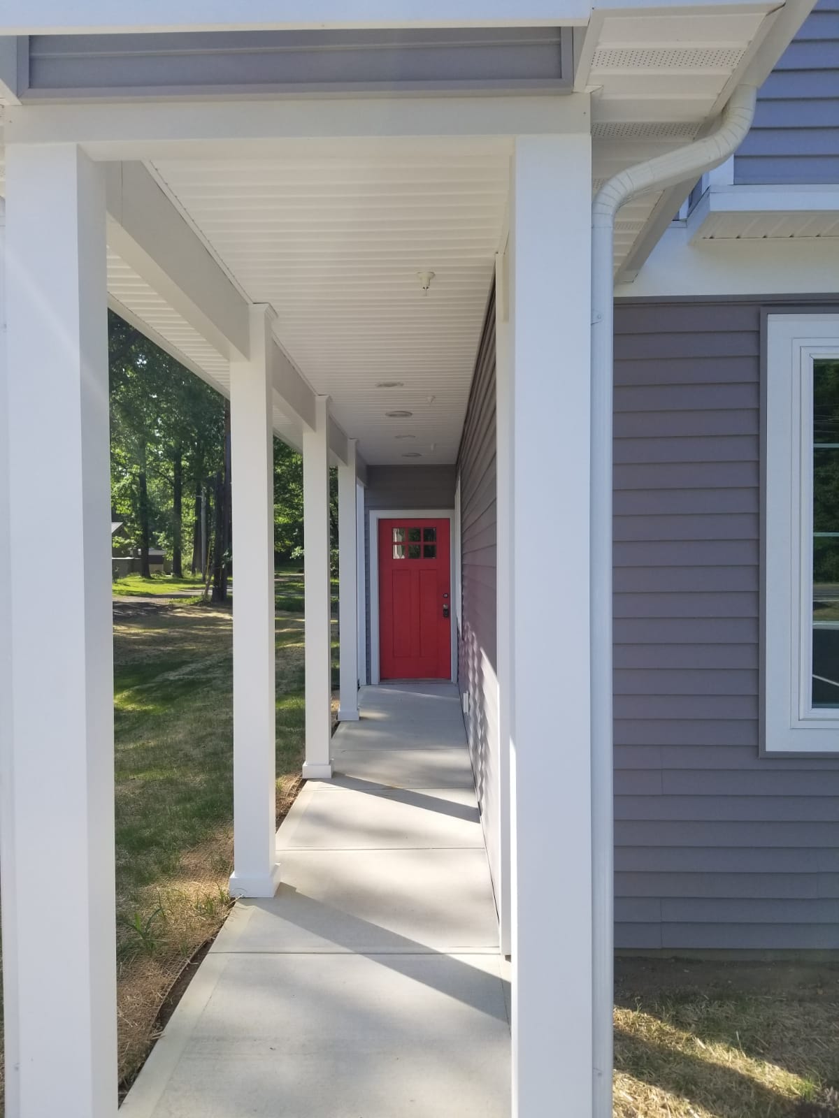 A covered walkway is shown leading to a red front door, framed by white columns. The pathway features concrete flooring, creating a clean and open approach. Sunlight filters through trees lining the exterior, casting gentle shadows along the path.
