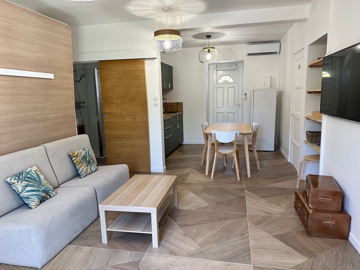 A brightly lit studio living area showcases a modern gray sofa adorned with tropical-patterned cushions. A light wooden coffee table is centered on a patterned floor. A dining table with white chairs sits against the wall, next to a kitchenette with sleek cabinets and a refrigerator.