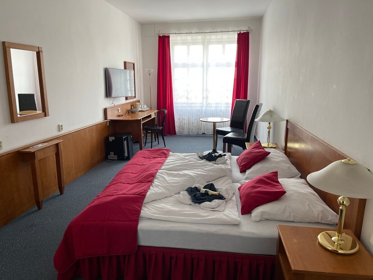 A spacious sleeping area features a large bed with a red coverlet and white pillows. A desk and chair are positioned next to a window, where natural light filters through sheer drapes. A television is mounted on the wall, and a full-length mirror is visible.
