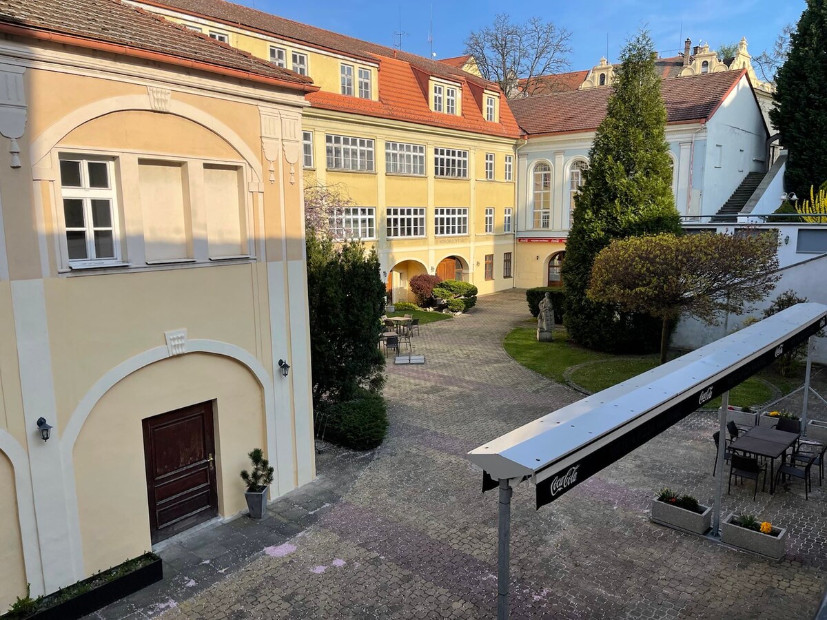 A serene courtyard is visible, surrounded by historical buildings with light-colored facades. Lush greenery is present with shrubs and trees, while outdoor seating areas are positioned under a covered structure. Pathways made of stone lead through the space, providing a peaceful atmosphere.