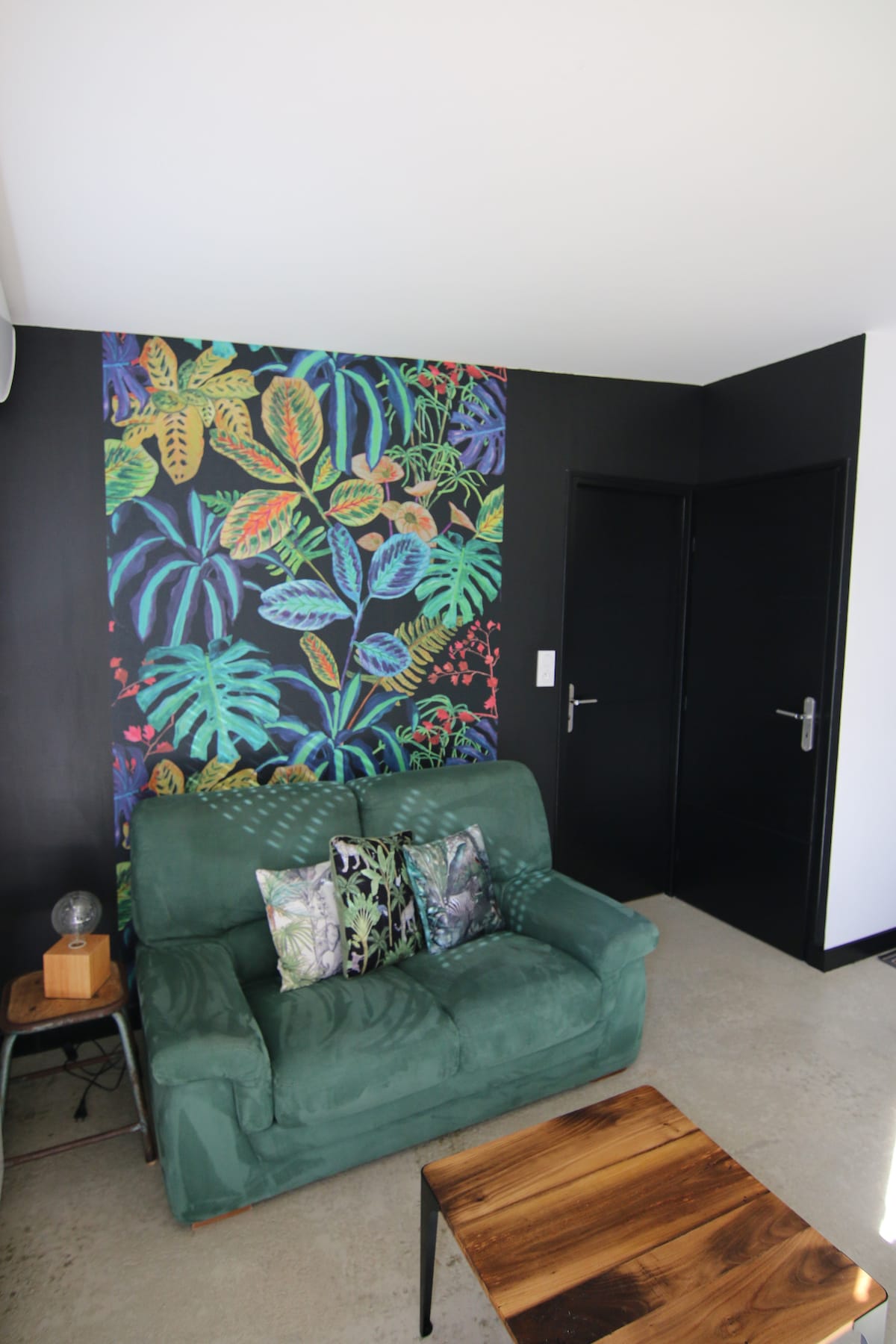 A cozy seating area features a green sofa adorned with patterned cushions. A wooden coffee table sits in front of the sofa, and a vibrant botanical mural decorates the wall behind, enhancing the space's connection to nature.