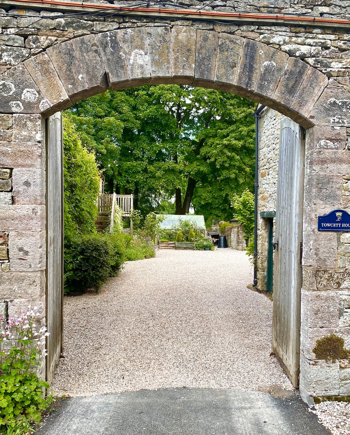 The entrance features a rustic stone archway, leading to a gravel pathway surrounded by lush greenery. Flanking the path are flowering plants and a hint of a greenhouse can be seen in the distance, suggesting a serene outdoor space.