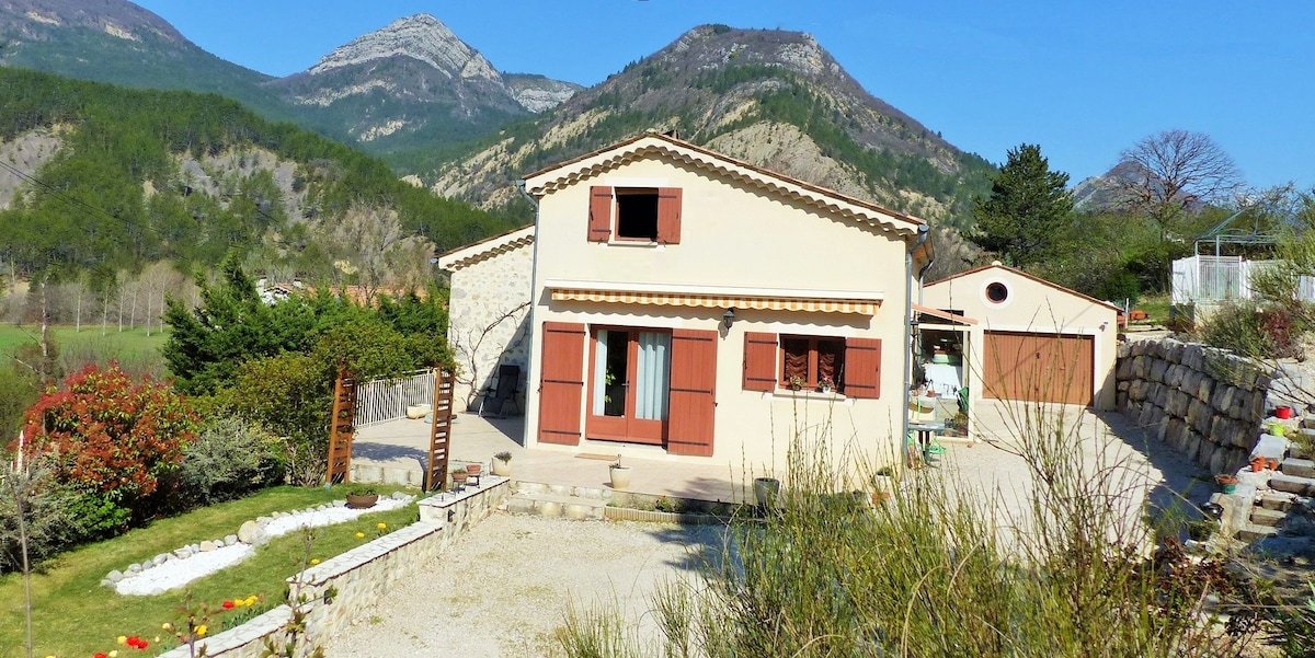 A two-story villa is set against a backdrop of rolling hills and mountains. The structure, featuring warm-toned stone and a light facade, includes multiple windows with wooden shutters. The surrounding landscape consists of trees and well-maintained garden areas, highlighting the property's spacious outdoor environment.