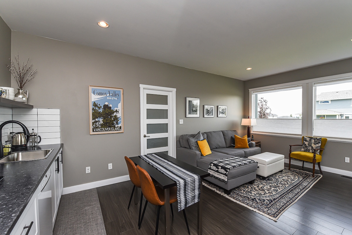 A light-filled living area features a sectional sofa, dining table, and decorative area rug. Large windows provide views of the outside, while a kitchenette with modern appliances can be seen along one wall. A framed artwork adds a touch of character to the space.