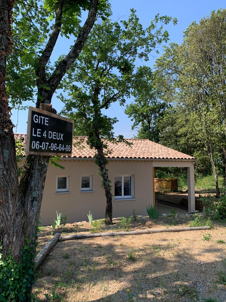 The exterior of the gîte features a light-colored façade, framed by trees and landscaped with gravel. A sign indicates the property name and contact information. A covered area is visible, providing shade, while several windows allow natural light to enter the interior.