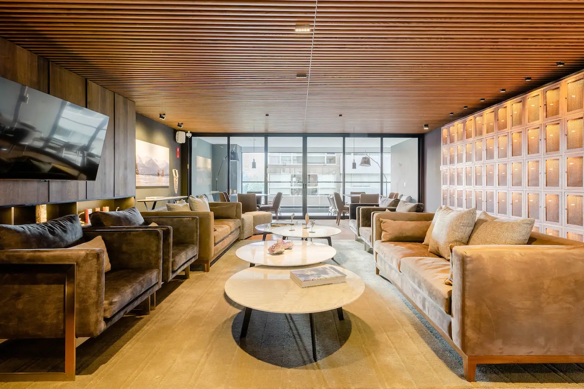 A modern lounge area features plush brown sofas arranged around circular tables. A large flat-screen television is mounted on the wall, and floor-to-ceiling windows allow natural light to fill the space. Soft lighting accentuates the wooden ceiling and the decorative backlit panels.