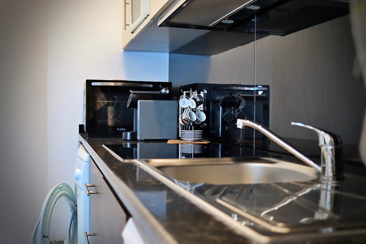 A modern kitchen is equipped with sleek cabinetry and a dark countertop. Essential appliances, including a microwave and coffee maker, are present. A dish rack holds several cups neatly. A stainless-steel sink is positioned beside a washing machine, contributing to the kitchen's functionality.