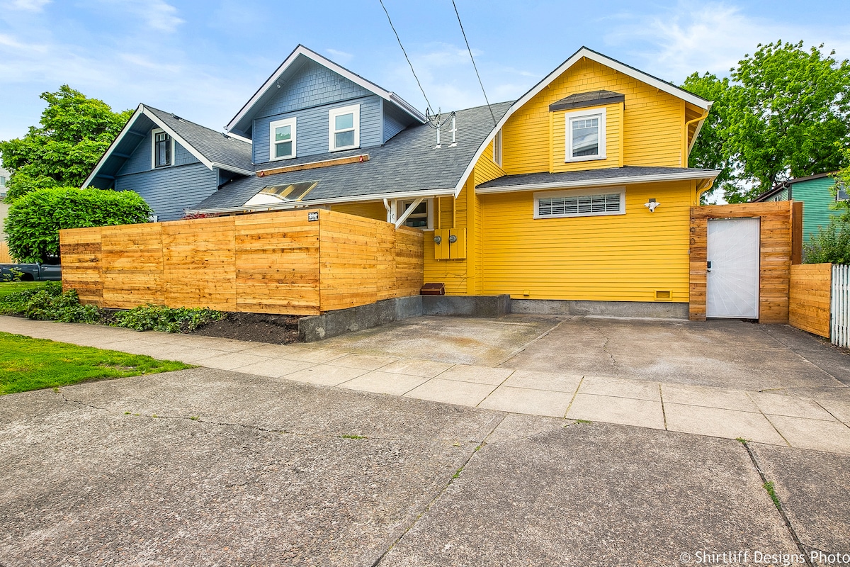 The exterior features a modern yellow house with a freshly painted facade. A wooden fence borders the property, and a paved driveway extends in front. The entrance gate is visible, providing easy access to off-street parking and the surrounding greenery.