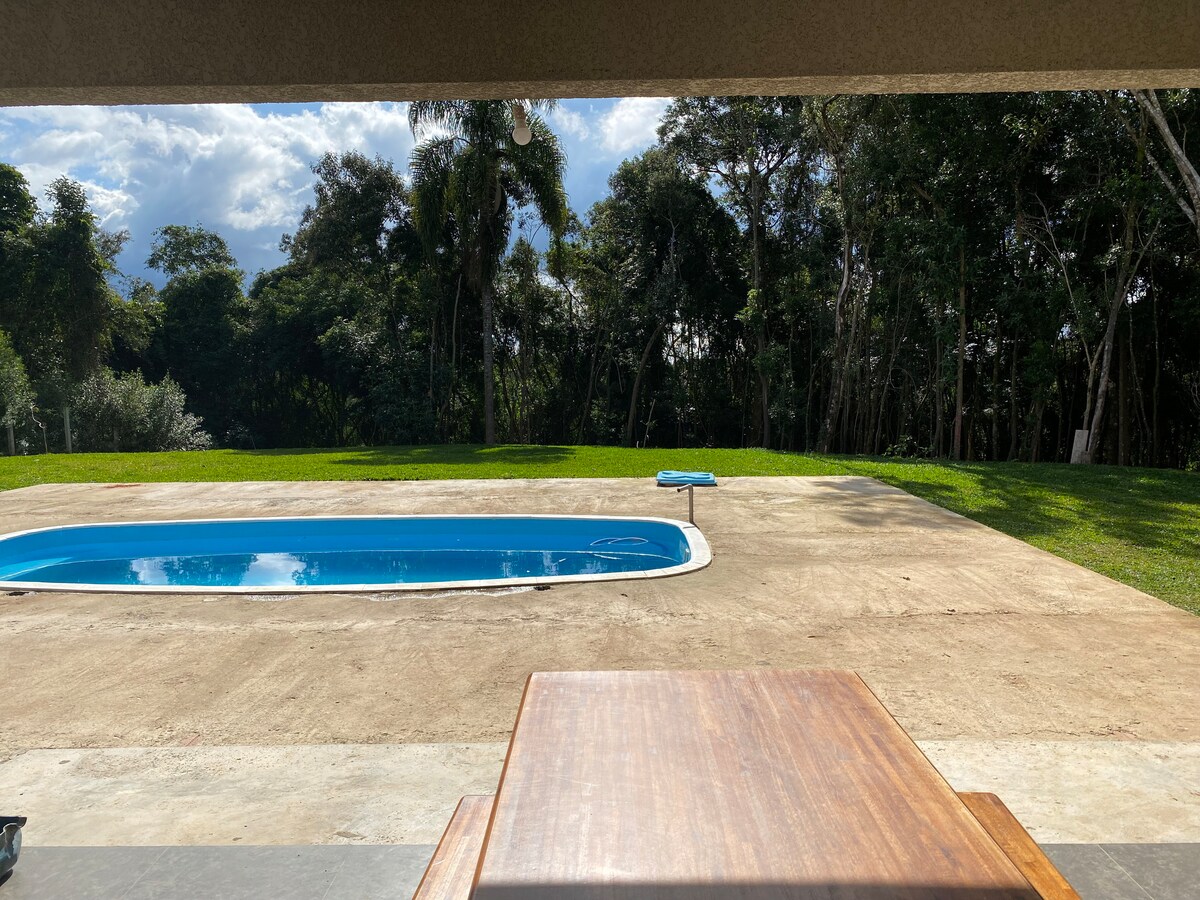 A clear view of a serene outdoor space showcases a rectangular swimming pool surrounded by a well-maintained lawn. Tall trees form a natural backdrop, enhancing the tranquility of the setting. A wooden table is positioned on a spacious concrete patio, providing a relaxing atmosphere.