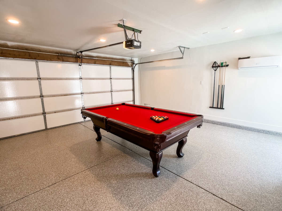 A recreational space is provided in the garage featuring a classic billiard table with a vibrant red felt surface at its center. The room is well-lit with overhead lighting and offers epoxy flooring, creating a clean and inviting environment.