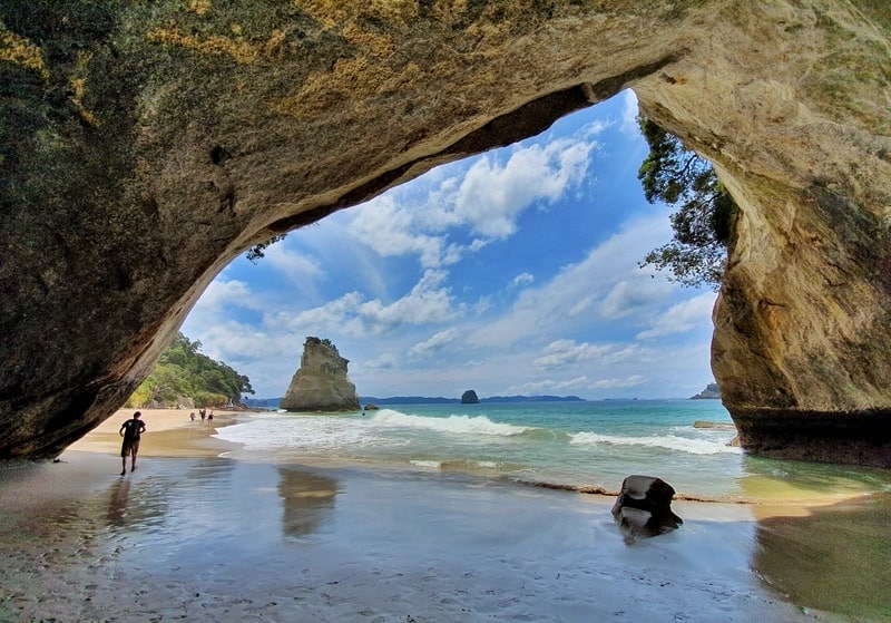 The image captures a coastal scene framed by a large rock arch, with soft sandy beach leading to gentle waves. Lush greenery is visible above the arch, while the horizon features distant landforms under a partly cloudy sky.