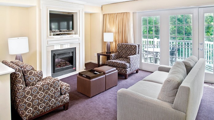 A spacious living area features a cozy seating arrangement with two patterned chairs and a neutral-toned sofa. A coffee table sits in the center, while an electric fireplace adds warmth. Large glass doors provide access to a balcony, allowing natural light to fill the room.