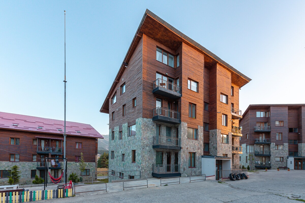 The exterior of the Villa Monte complex is depicted, featuring a multi-level building constructed with wood and stone materials. Balconies are visible on several floors, providing access to outdoor spaces. The surrounding area includes trees and open space, enhancing the natural ambiance.
