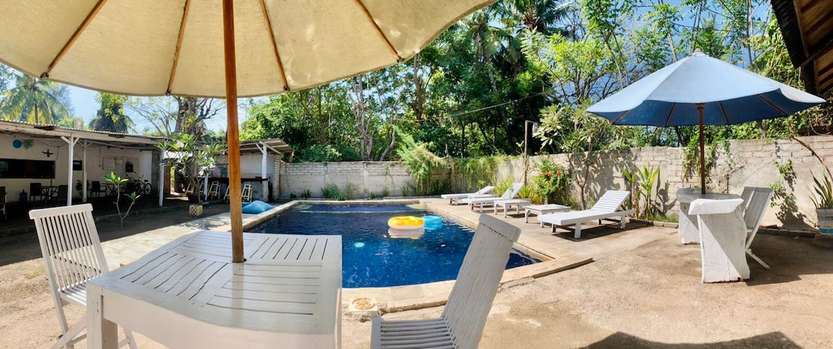 A swimming pool is surrounded by sun loungers and shaded areas. Bright umbrellas provide shelter beside the pool, while tropical plants create a natural backdrop. The clear water reflects the blue sky, inviting relaxation in a serene outdoor space.