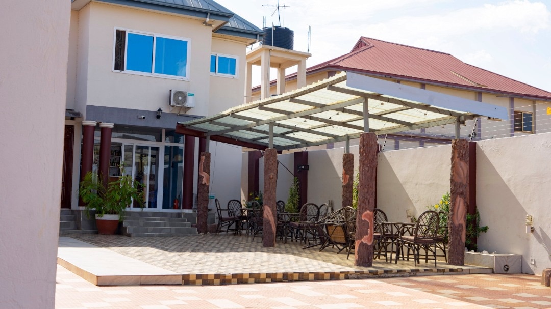 An outdoor seating area is highlighted, featuring several wicker chairs arranged under a glass canopy. The space is surrounded by stone walls, and decorative plants add a touch of greenery. The well-maintained patio is paved with square tiles, contributing to a welcoming environment.