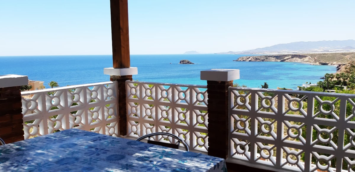 A spacious outdoor terrace provides a scenic view of the clear blue sea and distant coastline. A table with a patterned cover is set for dining, while decorative railings frame the picturesque landscape, creating an inviting atmosphere for relaxation.