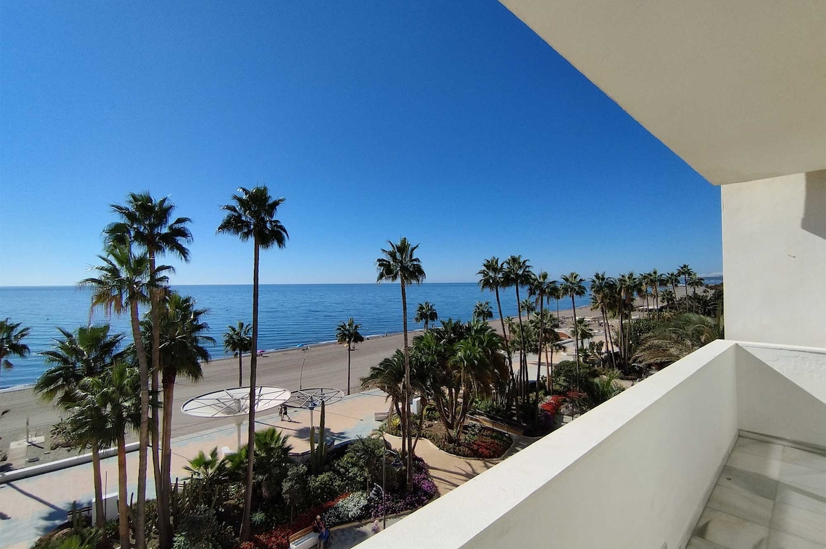 An expansive view overlooking the Mediterranean Sea is captured from the terrace. Lush palm trees line the coast, and a sandy beach stretches into the distance. The clear blue sky complements the tranquil waters, creating a serene coastal atmosphere.