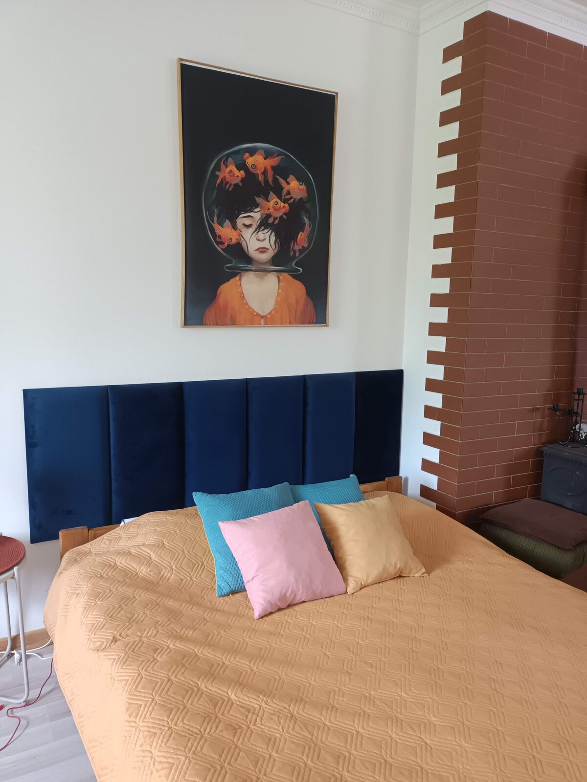 A well-furnished bedroom includes a spacious bed with an orange coverlet and colorful pillows in shades of blue and pink. A large framed artwork features a figure with a floral headpiece, positioned above the bed, while a brick wall adds texture to the room.
