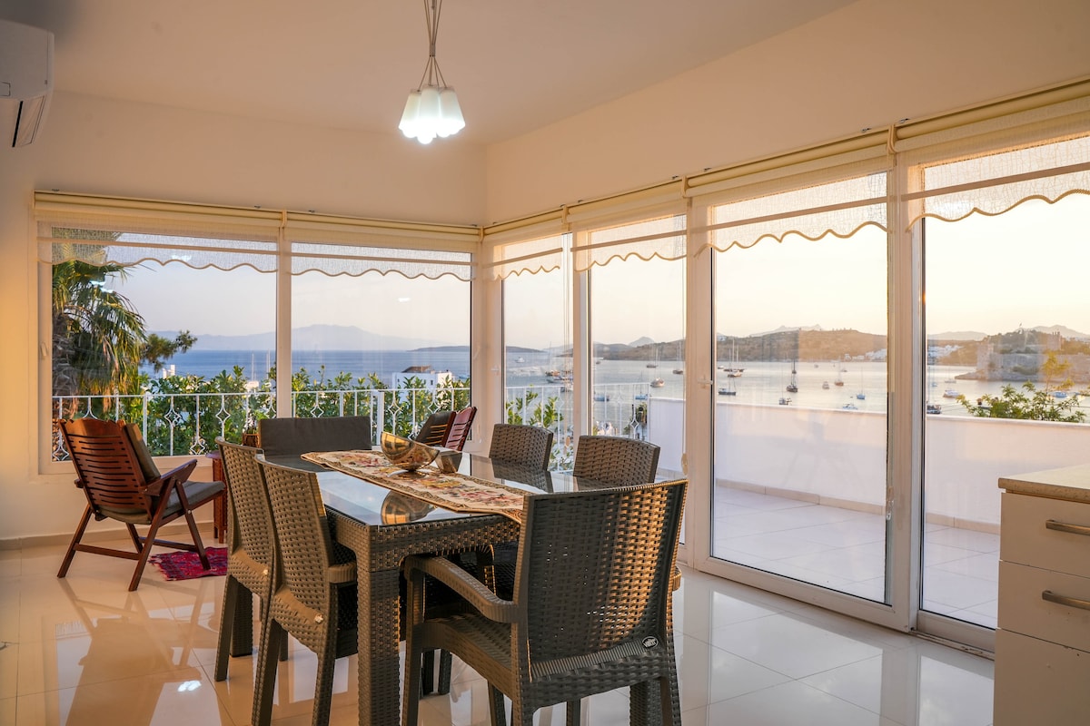 A bright dining area features a spacious table surrounded by modern wicker chairs, with panoramic windows showcasing a stunning sea view. Natural light floods the space, highlighting the clean lines and contemporary design. A cozy armchair is positioned nearby for relaxation.