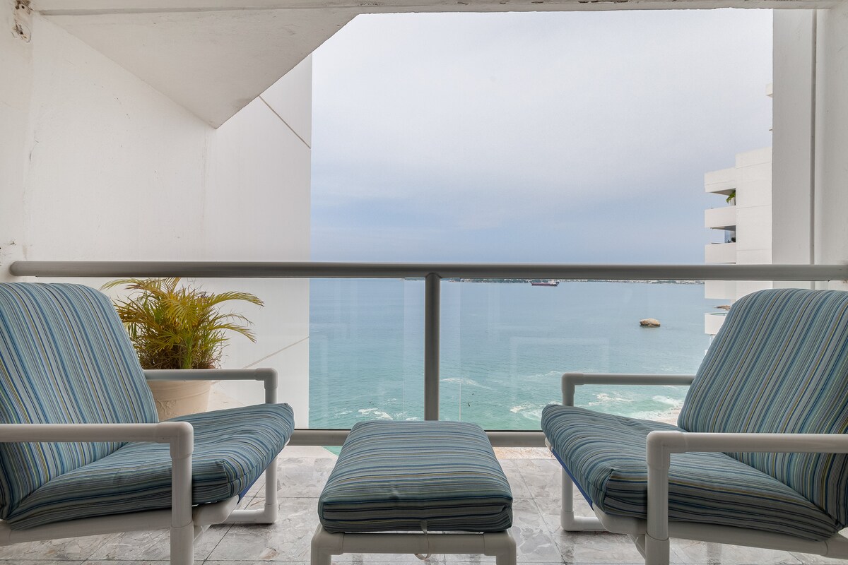 A balcony view features two striped chairs and a matching ottoman facing the ocean. The tranquil waters extend to the horizon, with subtle waves visible. A potted plant adds a touch of greenery, enhancing the coastal atmosphere.