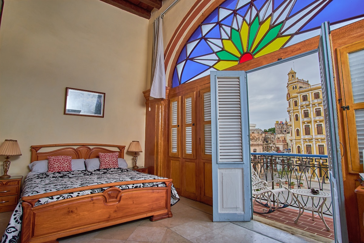 A spacious bedroom features a large wooden bed with a decorative black and white bedding set. Shuttered doors open to a private balcony overlooking the Plaza Vieja, revealing vibrant stained glass detailing and views of historic buildings.