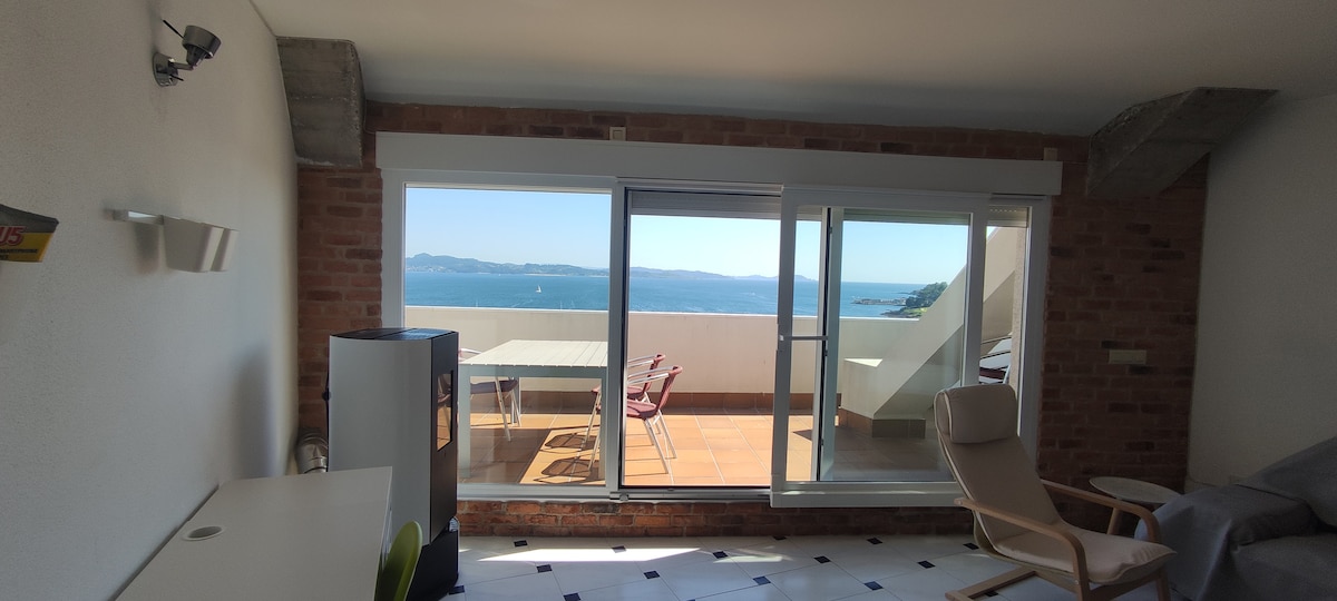 A spacious living area is characterized by large glass doors that open to a terrace with sea views. The room features a simple dining table with chairs and a comfortable armchair, while natural light fills the space through the windows, highlighting the exposed brick walls.