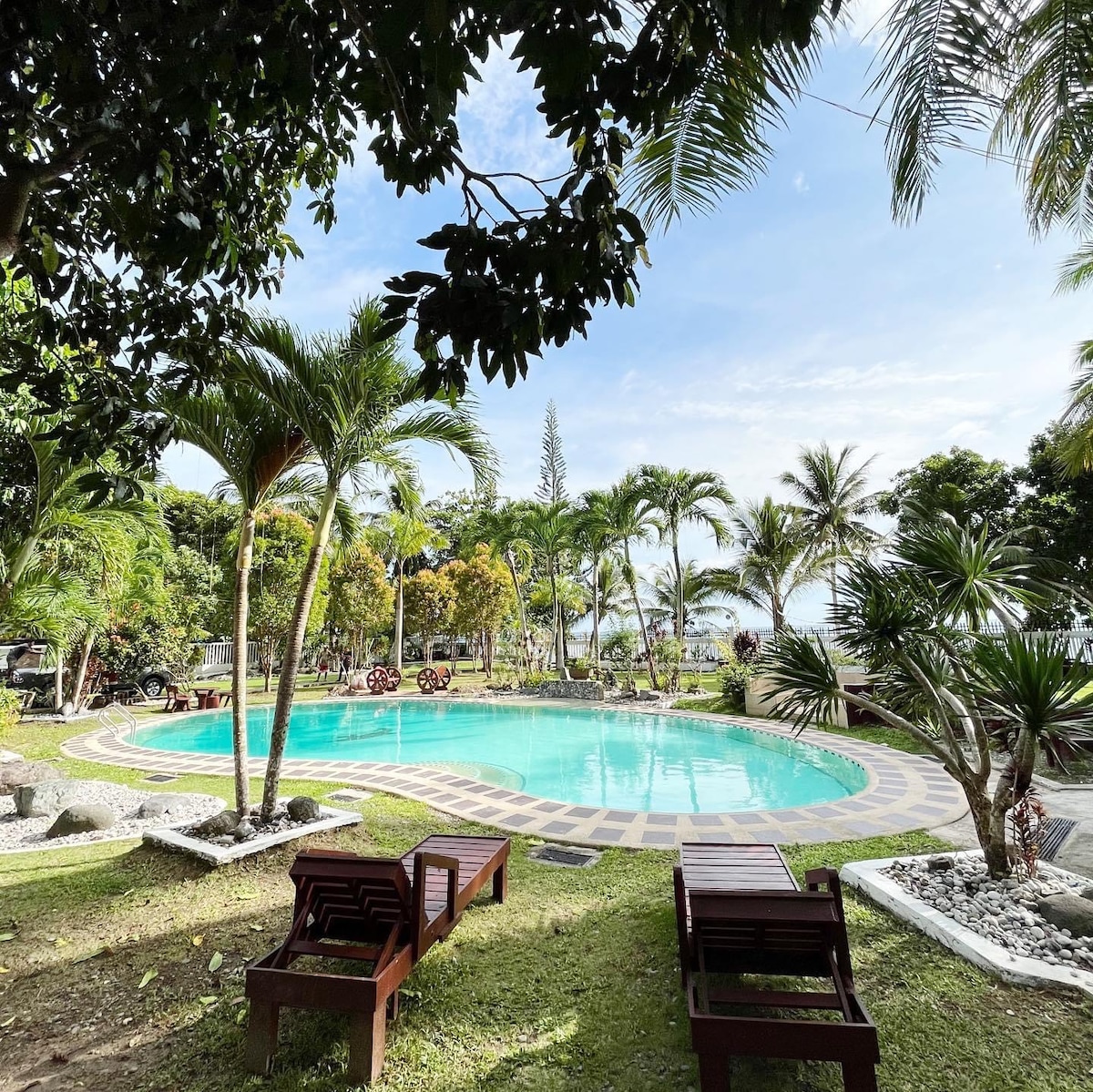 A tranquil swimming pool is surrounded by lush tropical plants and palm trees. Two wooden lounge chairs are positioned on the grass nearby, offering views of the clear water and landscaped gardens. The beach can be seen in the distance, completing the serene atmosphere.