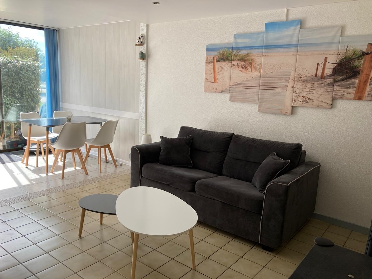 A living area is shown with a dark gray sofa and modern white coffee table. A canvas print depicting a sandy pathway is displayed on the wall. In the background, a dining area with a table and four chairs can be seen, adjacent to sliding glass doors that lead outside.