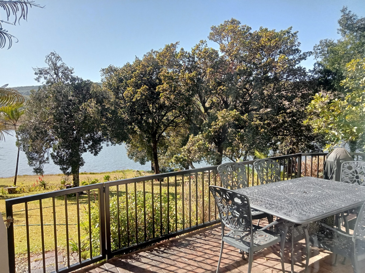 A patio area is shown featuring a black metal table surrounded by chairs. The space overlooks the lush green lawn leading to the tranquil water of the Da Gama dam, framed by tall trees and a clear blue sky.