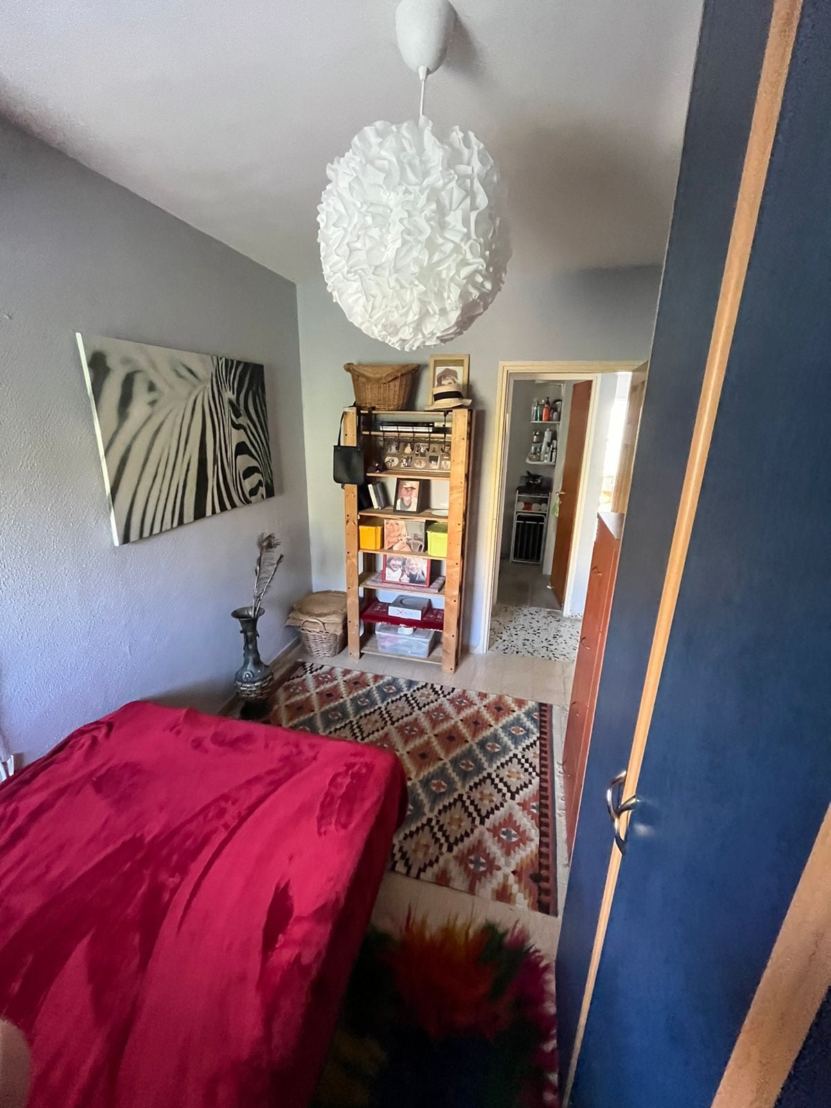 A cozy bedroom is presented, featuring a red blanket draping over a spacious bed. A textured rug lies on the floor, complementing the wood shelves filled with various items. Natural light enters through the doorway, illuminating the soft wall colors and decorative elements.
