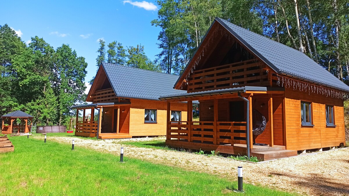 Two charming wooden cabins are positioned on a grassy area, surrounded by trees. Each cabin features a gabled roof and a spacious porch with wooden railings. A small gazebo can also be seen nearby, providing a shaded area for relaxation.