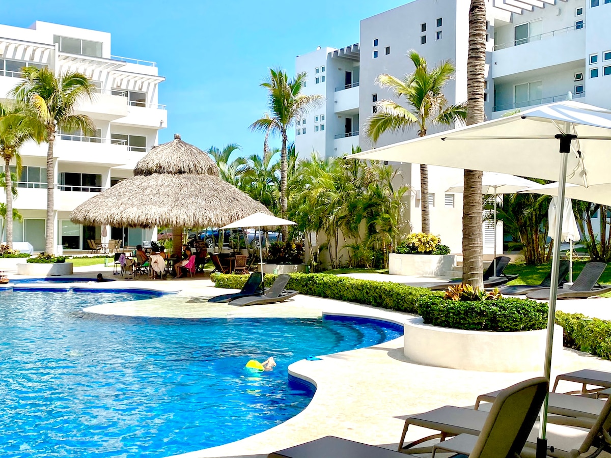 A swimming pool is framed by lush tropical plants, with lounge chairs arranged nearby. A thatched-roof palapa offers shaded seating for guests. The clear blue water reflects the surrounding bright skies and modern white buildings of the complex.