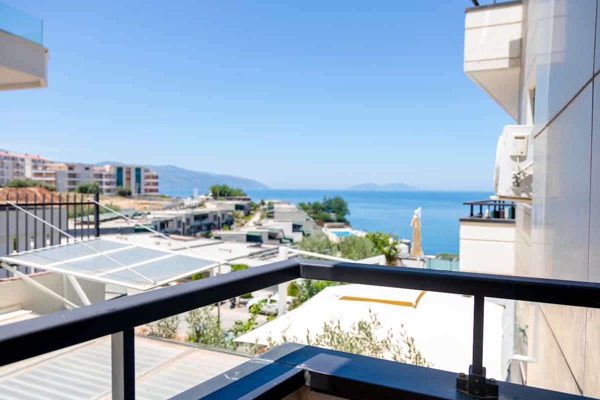 A balcony view features a clear horizon of the sea, framed by a railing. The landscape includes a mix of buildings and greenery in the foreground, with distant mountains visible against a blue sky. The tranquil scene suggests an inviting atmosphere for relaxation.