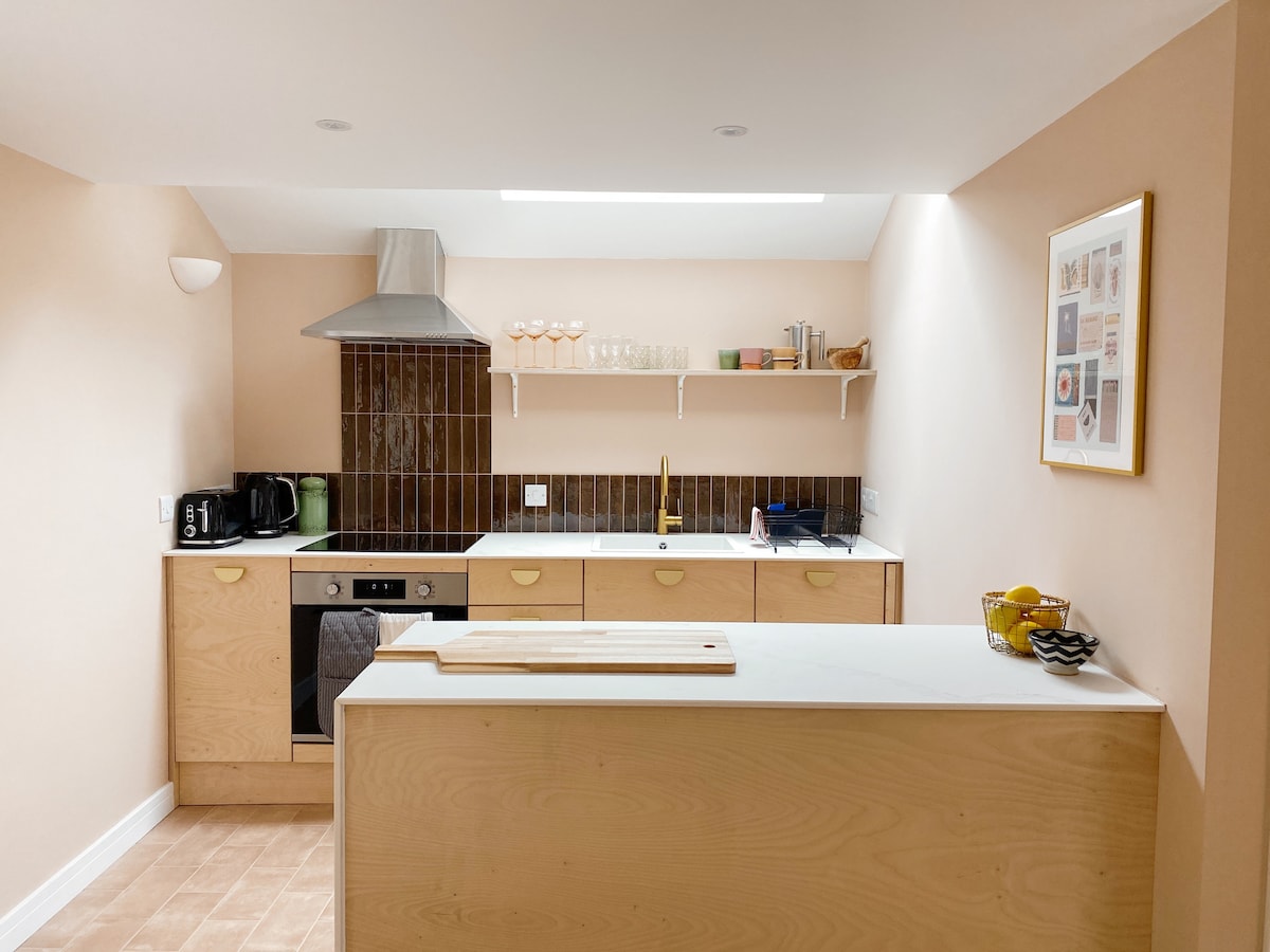 A modern kitchen is shown with beige walls and a wood-topped island. Features include a stainless steel range hood, minimalist cabinetry, and dark tiled backsplash. Kitchenware is neatly arranged on open shelves, with a bowl of fresh fruit on the counter and a variety of appliances visible.