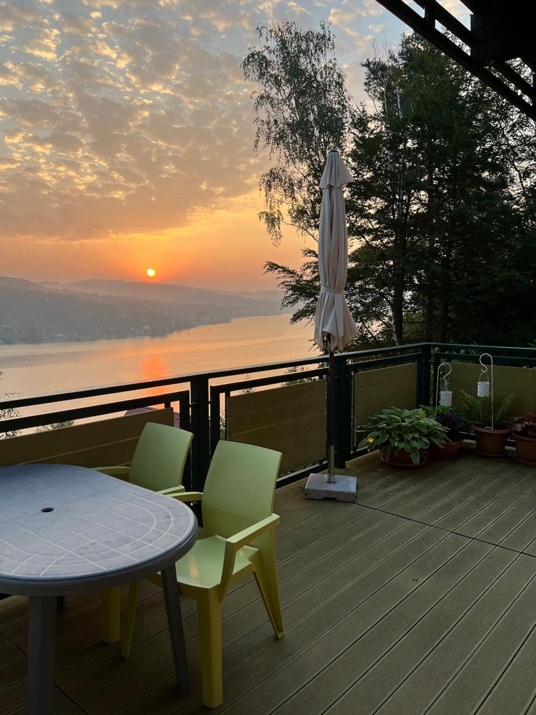 A tranquil outdoor deck is depicted, featuring a round table with four chairs and lush potted plants. In the background, a stunning sunset casts warm hues over the calm waters of Lake Domasa, framed by trees and a clear sky.