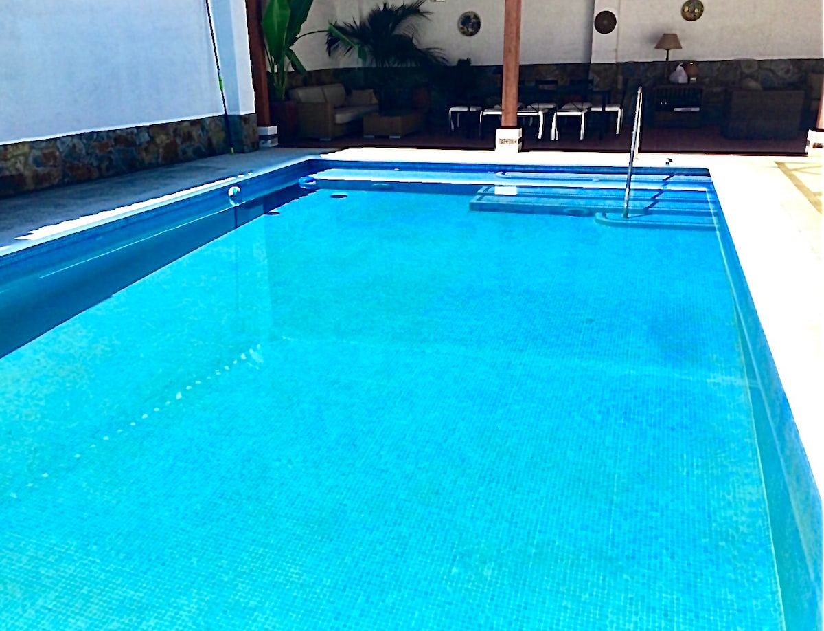 A private swimming pool with clear blue water is visible under natural light. Comfortable outdoor seating is arranged nearby, providing areas for relaxation. The pool is designed with tiles and features steps leading into the water.