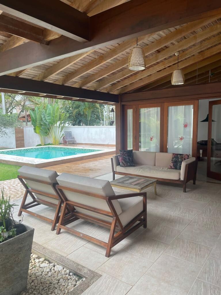 An outdoor seating area is presented, featuring two wooden chairs and a small table. The space is surrounded by greenery and offers a view of a swimming pool. Natural light filters through the structure's open design, enhancing the relaxing atmosphere.