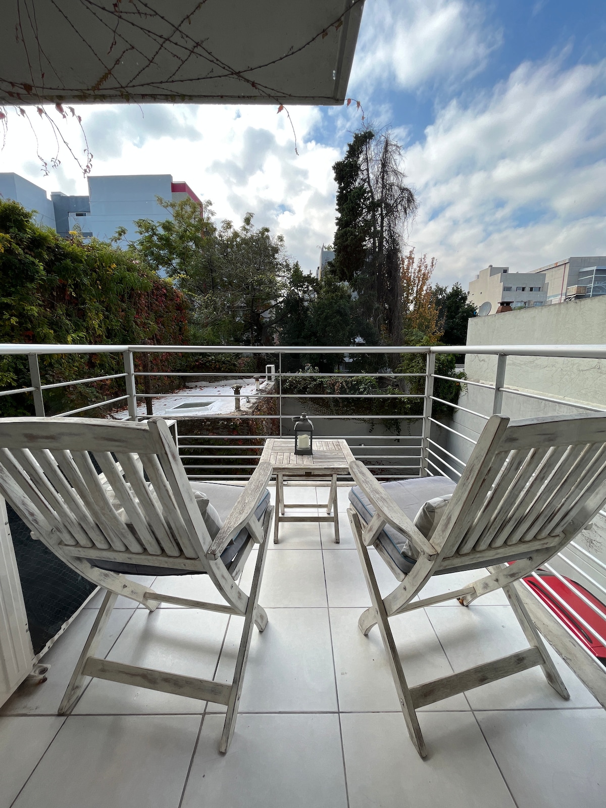 A balcony area is featured with two wooden chairs positioned around a small table. Green foliage and trees are visible beyond the railing, creating a tranquil outdoor setting. The sky is partly cloudy, adding to the brightness of the space.